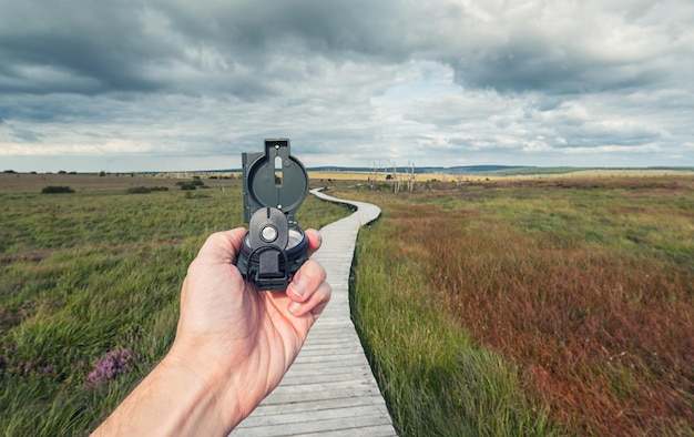 Mannelijke reiziger houdt een kompas vast op de achtergrond van een moeraslandschap met houten pad en bewolkte hemel, zicht op de hand, pov.