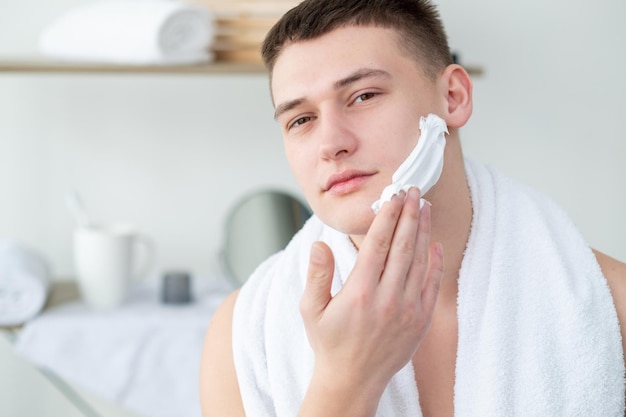 Mannelijke scheerroutine Huidverzorging cosmetologie Dagelijkse scheerbeurt Zelfverzekerde shirtloze man met witte handdoek op schouders schuim aanbrengen op gladde perfecte huid in lichte badkamer