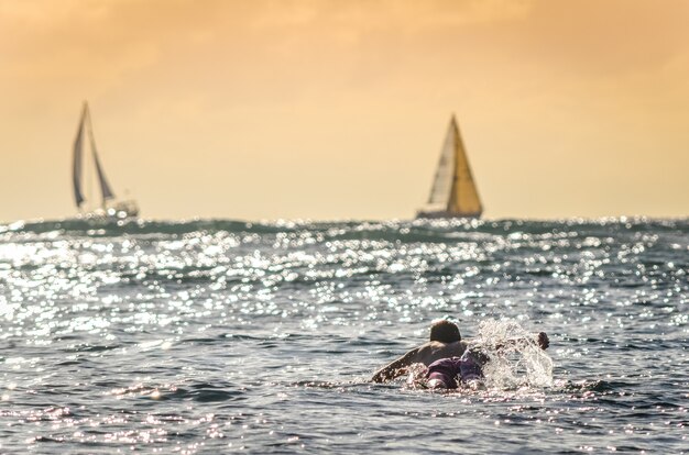 Mannelijke surfer die uit bij zonsondergang in hawaï met zeilboten op de achtergrond paddelen