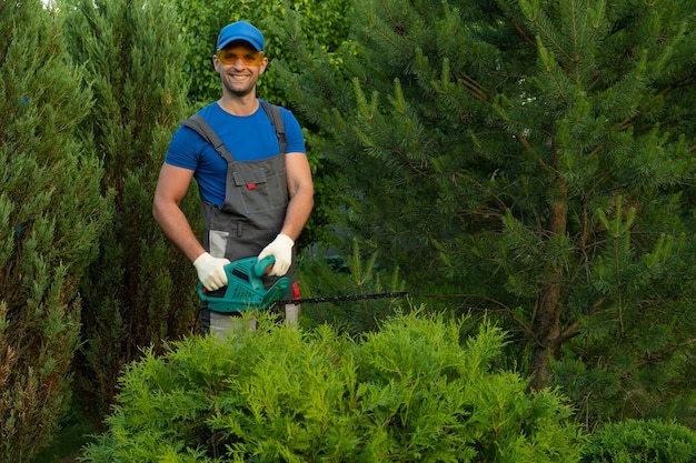 Mannelijke tuinman in een beschermend pak snoeit struiken met een elektrische trimmer