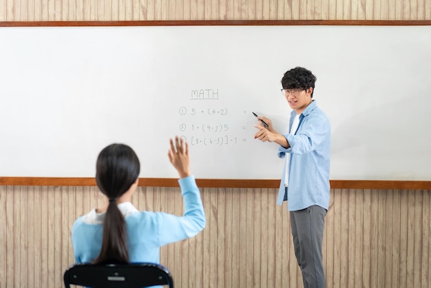 Foto mannelijke tutor die voor het whiteboard staat, wijst naar de student om een vraag te stellen en jonge studenten steken de handen in de lucht om een vraag te beantwoorden tijdens het leren in de klas