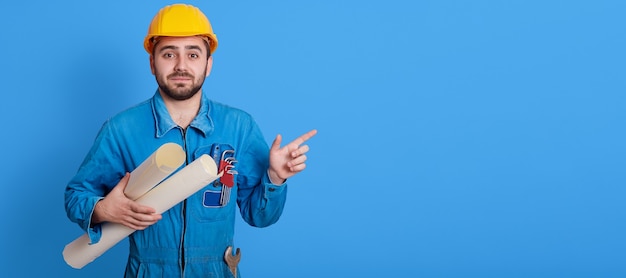 Mannelijke werknemer blauwdrukken houden en opzij wijzen met wijsvinger, ongeschoren ingenieur gele helm en blauw uniform dragen