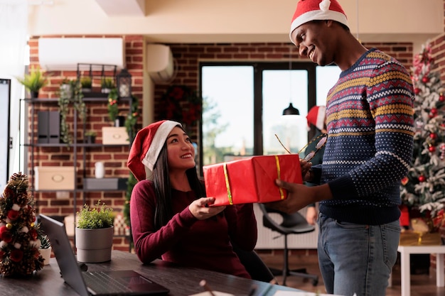 Mannelijke werknemer die geschenkdoos geeft aan aziatische vrouw in feestelijk kantoor versierd met kerstboom en ornamenten. Man viert wintervakantieseizoen met cadeautjes op zakelijke werkplek.