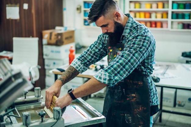 Mannelijke werknemer die inkt op het frame drukt tijdens het gebruik van de drukmachine in een werkplaats