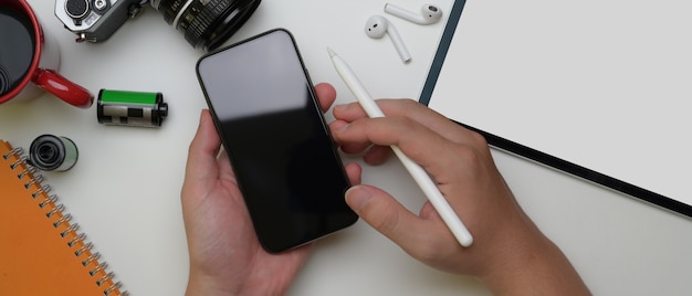 Mannelijke werknemer die smartphone boven wit bureau met tablet, camera en andere levering gebruiken