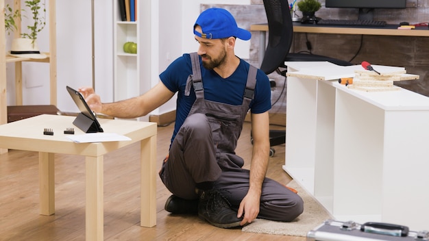 Mannelijke werknemer in overall assembleert nieuwe witte meubels in nieuwe huiseigenaren. Man leest instructie op laptop.