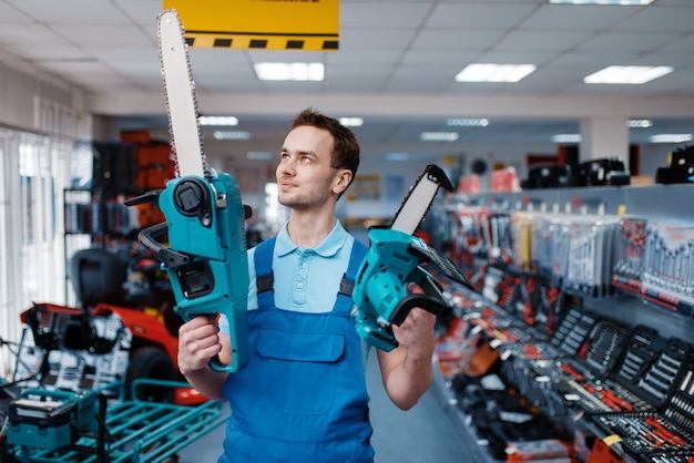 Mannelijke werknemer in uniform houdt grote en kleine kettingzagen in gereedschapsopslag. keuze uit professionele apparatuur in ijzerhandel, instrumentensupermarkt