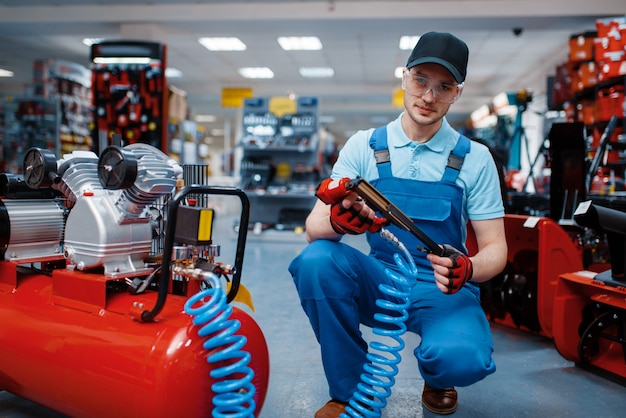 Mannelijke werknemer in uniforme poses met pneumatische spijkermaker in gereedschapsopslag. keuze uit professionele apparatuur in ijzerhandel, instrumentensupermarkt