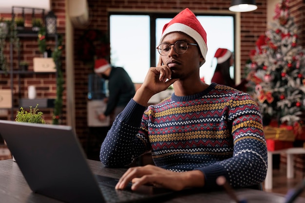 Mannelijke werknemer met kerstmuts met behulp van laptop in feestelijk ingerichte werkplek. Werken aan project in kantoor met kerstboom en decoraties, seizoensgebonden vakantie ornamenten.