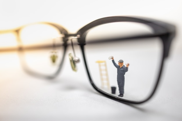 Foto mannelijke werknemer miniatuur figuur veeg en maak een vuile leesbril met emmer en ladder op witte tafel schoon.