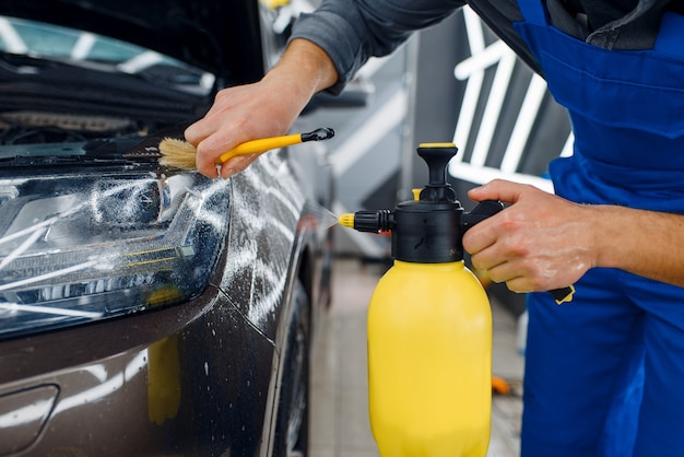 Mannelijke werknemer reinigt auto-oppervlak met spray en borstel, voorbereiding voor het aanbrengen van beschermfolie. Installatie van coating die de autolak tegen krassen beschermt. Voertuig in garage