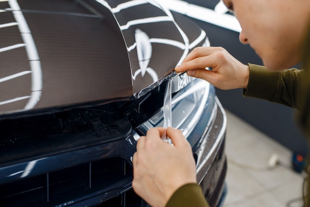 Mannelijke werknemer snijdt transparante beschermfolie op de motorkap. Installatie van coating die de autolak tegen krassen beschermt. Nieuw voertuig in garage, afstemmingsprocedure