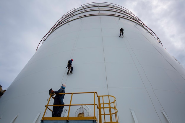 Mannelijke werknemers controleren de kabel naar beneden, de toegang tot de tank, de inspectie van de dikte van de schaal van de schaal van de opslagtank, veiligheidswerkzaamheden op hoogte.