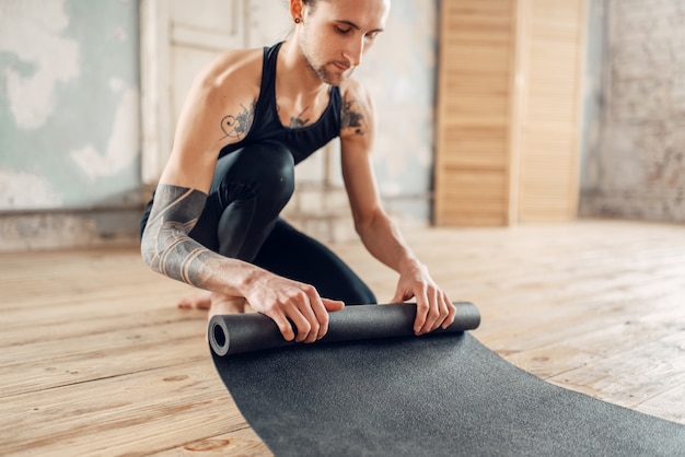 Mannelijke yoga met tatoeage bij de hand bereidt mat voor op training in de sportschool met grunge interieur.