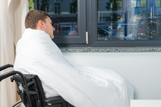 Mannelijke zieke patiënt zit bij het raam op rolstoel bedekt met quilt in ziekenhuisafdeling. zorgconcept