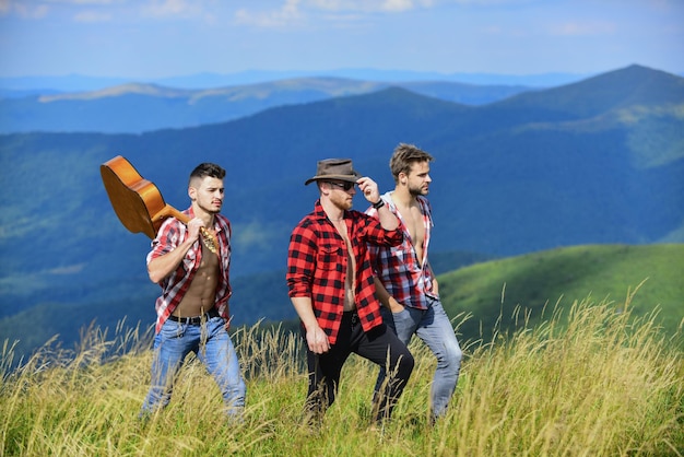 Mannen met gitaar wandelen op zonnige dag. Wandelen met vrienden. Lange weg. Avonturiers ploeg. Toeristen wandelen concept. Groep jongeren in geruite overhemden die samen op de top van de berg lopen.