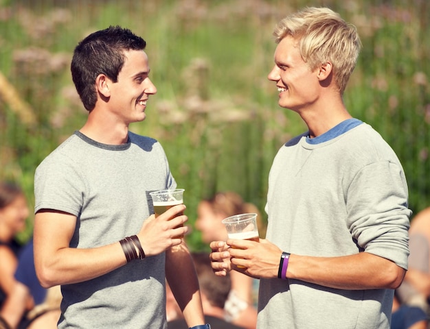 Mannen praten en drinken in de zomer met bier alcohol en geluk op een openlucht muziekfestival of evenement Gelukkig gesprek en vrienden in het park voor concertviering en samen drinken in het bos