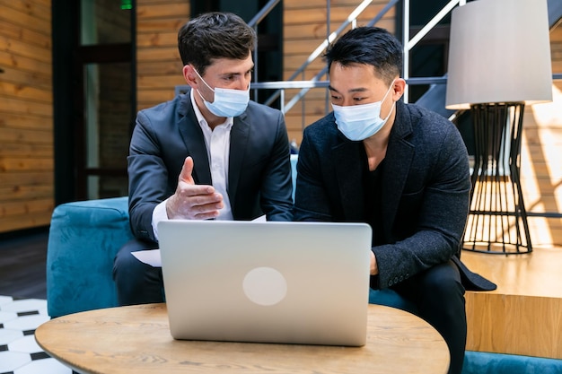 Mannen zitten op de bank en bespreken gezamenlijke zaken op een laptop met een gezichtsmasker op