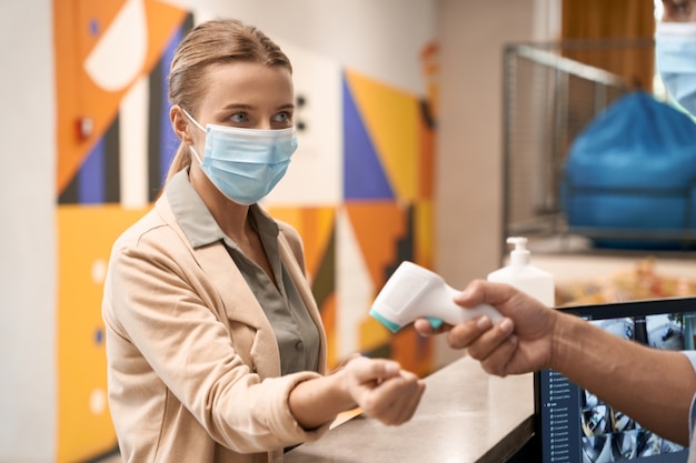 Mannenhand die de lichaamstemperatuur meet van een jonge zakenvrouw met een medisch masker of een vrouwelijk kantoor