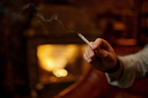 Mannenhand houdt een sigaret, rijk kantoor interieur met open haard. Tabaksrookcultuur, specifieke smaak. Roker vrije tijd thuis