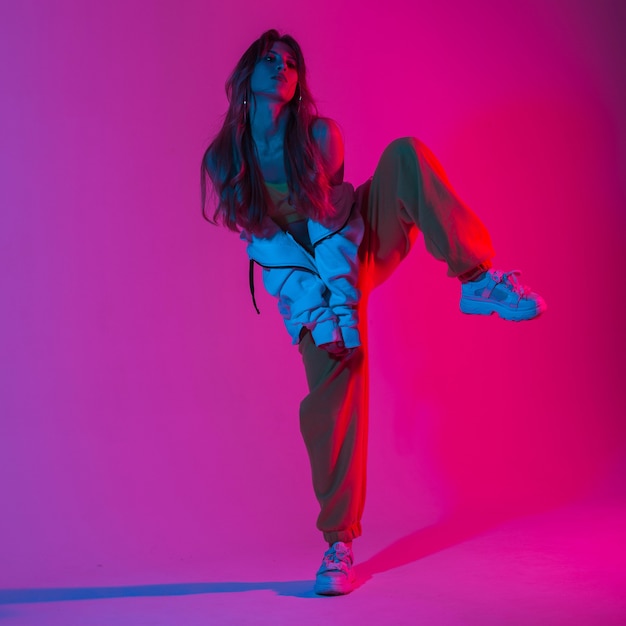 Mannequin aantrekkelijke vrouw in een stijlvol jeugdsportpak in trendy sneakers staat op één been in de studio met felroze licht. Meisjesdanser die op de achtergrond van een gekleurde studio dansen.
