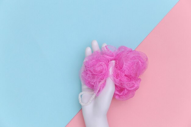 Mannequin hand holds bath sponge on blue-pink pastel background. Top view