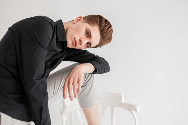 Mannequin knappe jongeman met trendy kapsel in modieus zwart shirt in stijlvolle broek staat in de buurt van stoel op witte achtergrond in studio. Knappe elegante kerel poseren in de buurt van muur binnenshuis.