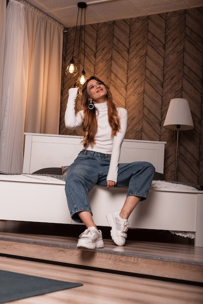 Mannequin van een chique jonge vrouw in een witte stijlvolle top in modieuze spijkerbroek poseren op het bed in de slaapkamer. Roodharig meisje in trendy kleding rust in de kamer met een modern interieur.