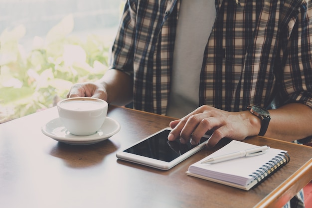 Mannetje dat een slimme telefoon met koffiekop op houten lijst gebruikt.