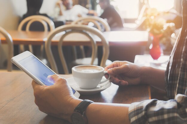 Mannetje dat een slimme telefoon met koffiekop op houten lijst gebruikt.