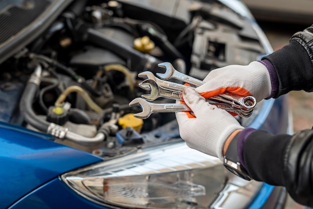 La mano della meccanica guantata dell'uomo tiene gli attrezzi dell'auto vicino a un'auto verniciata con un cofano aperto concetto di strumenti concetto di riparazione auto