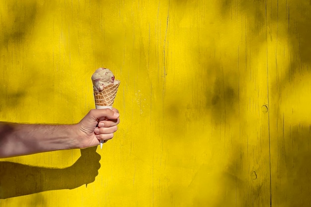 Photo mans hand holding an ice-cream in the cone over the bright yellow wall on a sunny summer day