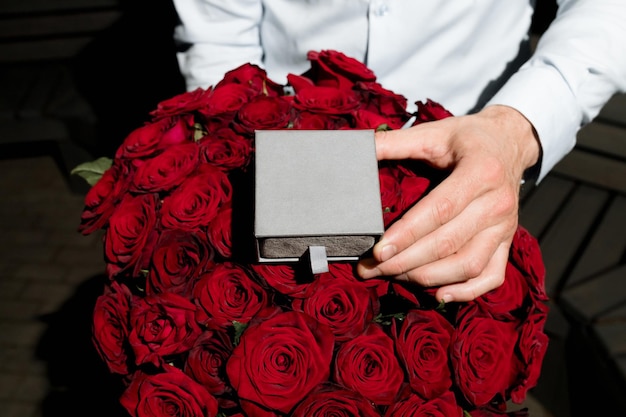 a mans hand holds a gray box with rings on a background of red roses an offer of marriage