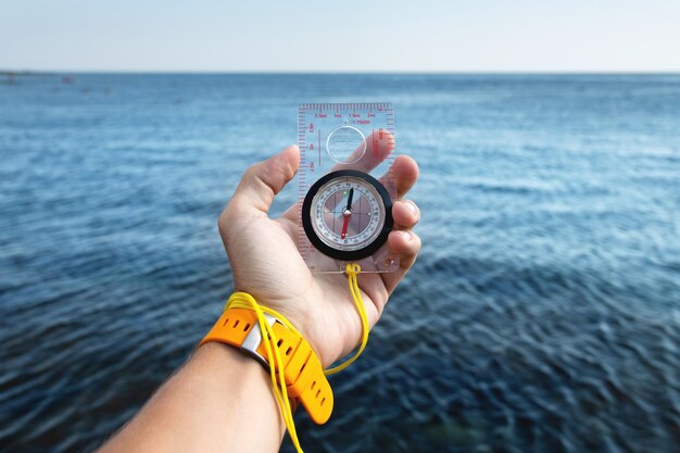 A mans hand with a wristwatch bracelet holds a magnetic compass against the background of the sea