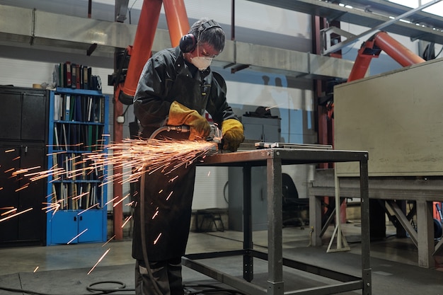 Manual worker in mask and ear protectors standing at high metal\
table and cutting metal with rotary tool in industrial shop