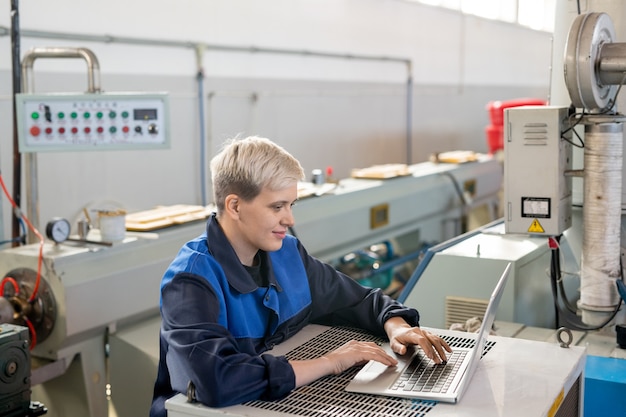 Manual worker in mask and ear protectors standing at high metal table and cutting metal with rotary tool in industrial shop