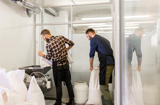 manufacture, business and people concept - men with bags weighing and pouring malt to mill at craft brewery or beer plant