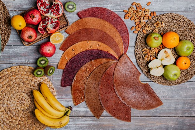 Many colorful fruit leathers with fresh fruits on the wooden table. Round fruit leather. Healthy food. Apples, bananas, nuts, lime, kiwifruits, pomegranate top view.