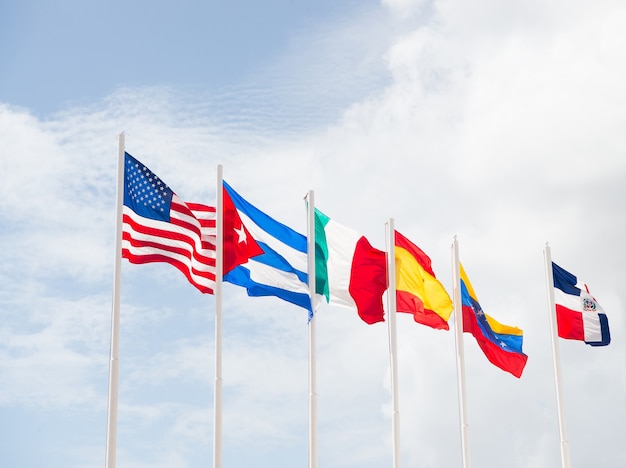 Photo many colorful national flags of different countries on masts outdoor on blue cloudy sky background, symbols of usa cuba ireland spain venezuela and dominican republic