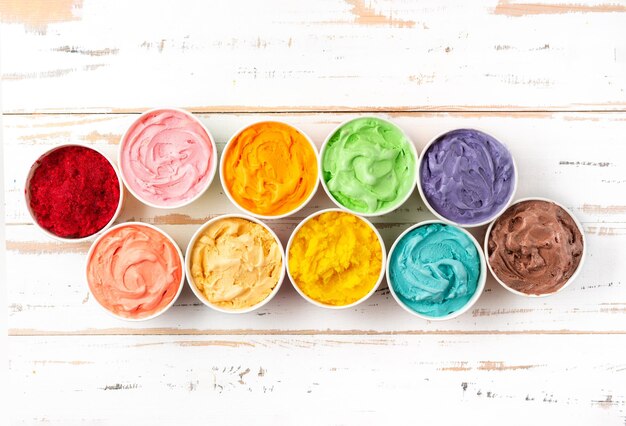 Many cups with colorful rainbow ice cream on white wooden background Top view