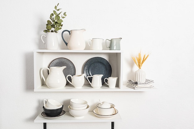 Many different jugs under water and milk are on the kitchen open shelves bowls and plates Beautiful white interior view of the kitchen shelves