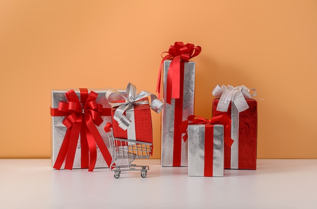 Many Gift Box with Red ribbon bow and shopping cart or trolley on white table and pastel orange wall