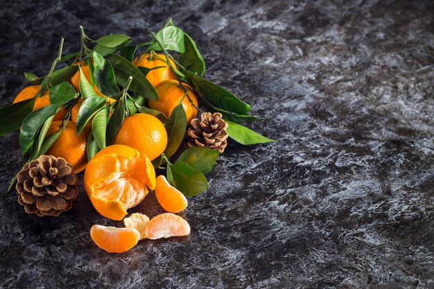 Many orange tangerines with green leaves on dark background with copy space. Peeled mandarin slices and cones