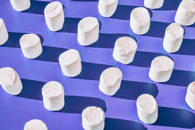 Many rows of white marshmallows cylindrical form lies on blue background. Pattern of unhealthy junky food. Sweets pattern