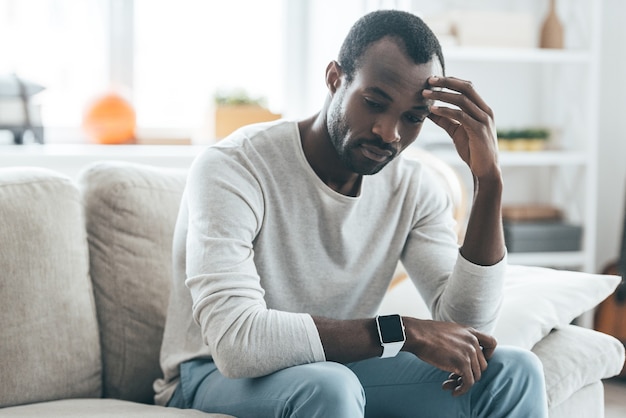 Many things to think about. Handsome young African man touching head with hand and looking uncertain while sitting on the sofa at home