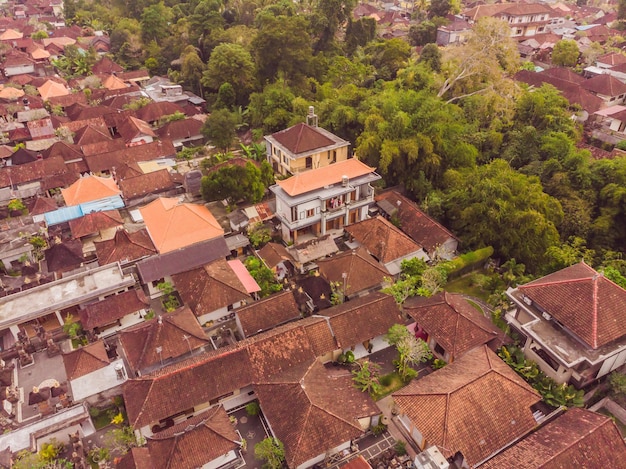 Many villas with brownorange shingle roofs between tropical trees on the sky background in Ubud on Bali Sun is shining onto them Aerial horizontal photo