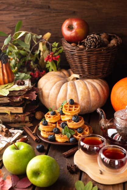 Many waffles are stacked on a plate, decorated with mint leaves and blueberries. Tea in a glass teapot and two small bowls.