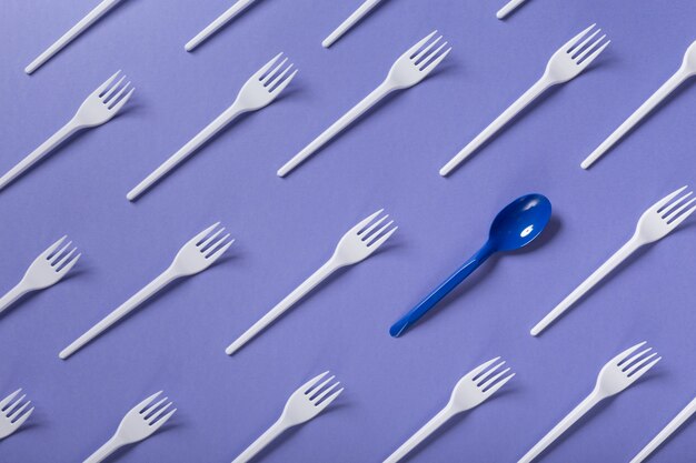 Many white plasic forks and one blue spoon on violet background with copy space, top view. Be brave and different, leadership concept