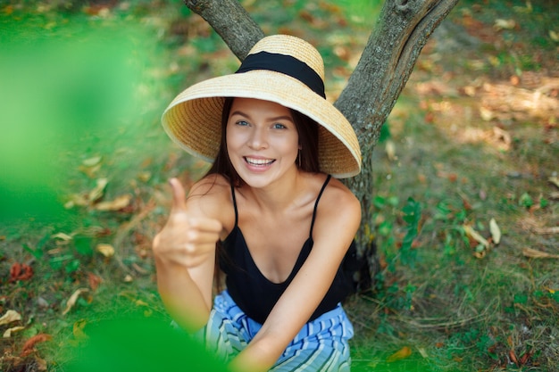 Mao Hainan Bamboo Conical Hat. Girl vegan, tourist, sitting under the tree on the grass, resting and showing class, hand gesture, finger up. A wide snow-white smile.