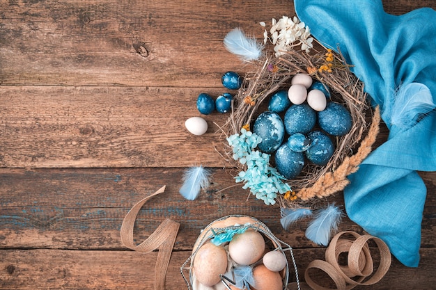 Фото Мраморно-синие пасхальные яйца в плетеном гнезде с перьями и цветами рядом с синей салфеткой и